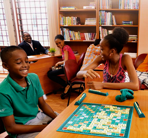 smiling children play a board game on a table in the foreground, while two adults look over from a court-related meeting in the background