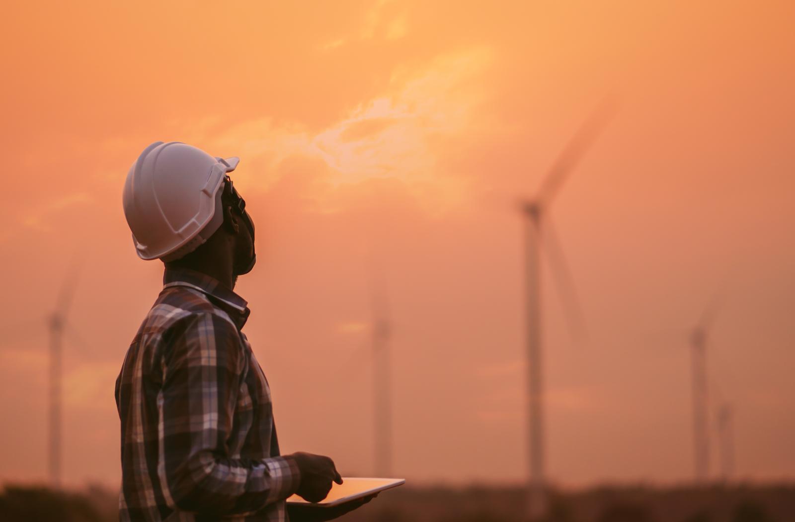 An engineer wearing a white hard hat with wind turbines behind him