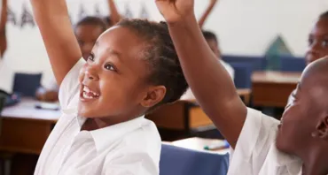 Young children at desks in classroom with their hands raised